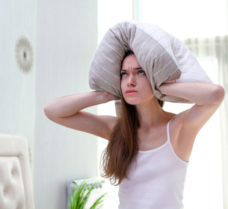 Angry woman suffering and disturbed by noisy neighbors and covering her ears with pillow while trying to falling sleep in bed at home in early morning.